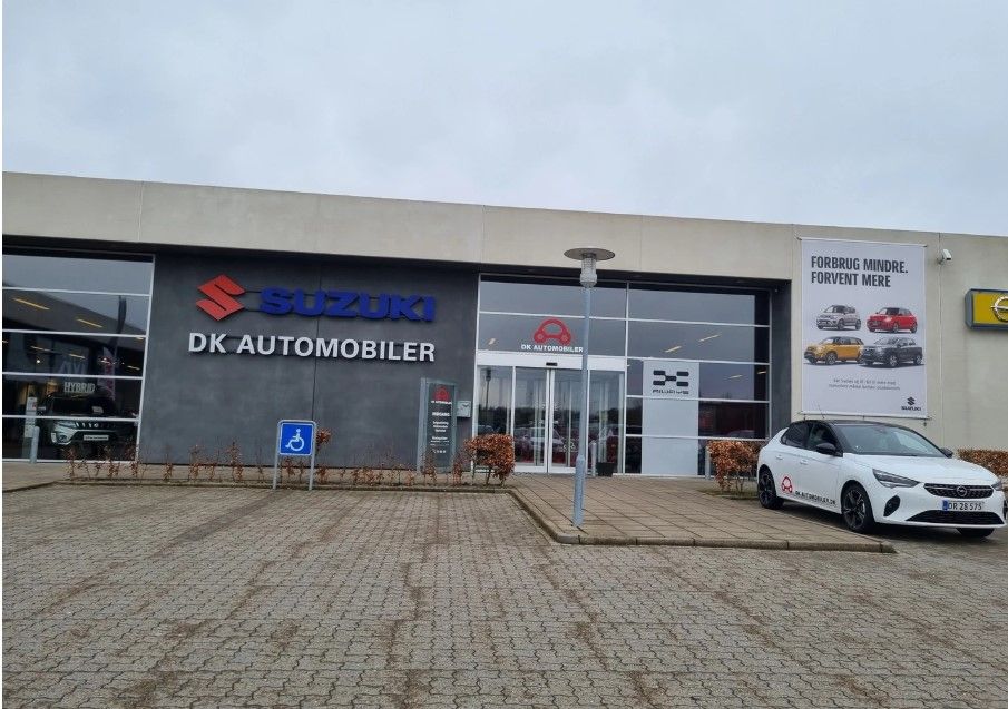 Suzuki DK Automobiler Vejle, med moderne showroom, en hvid bil parkeret og en banner med teksten Forbrug mindre, forvent mere