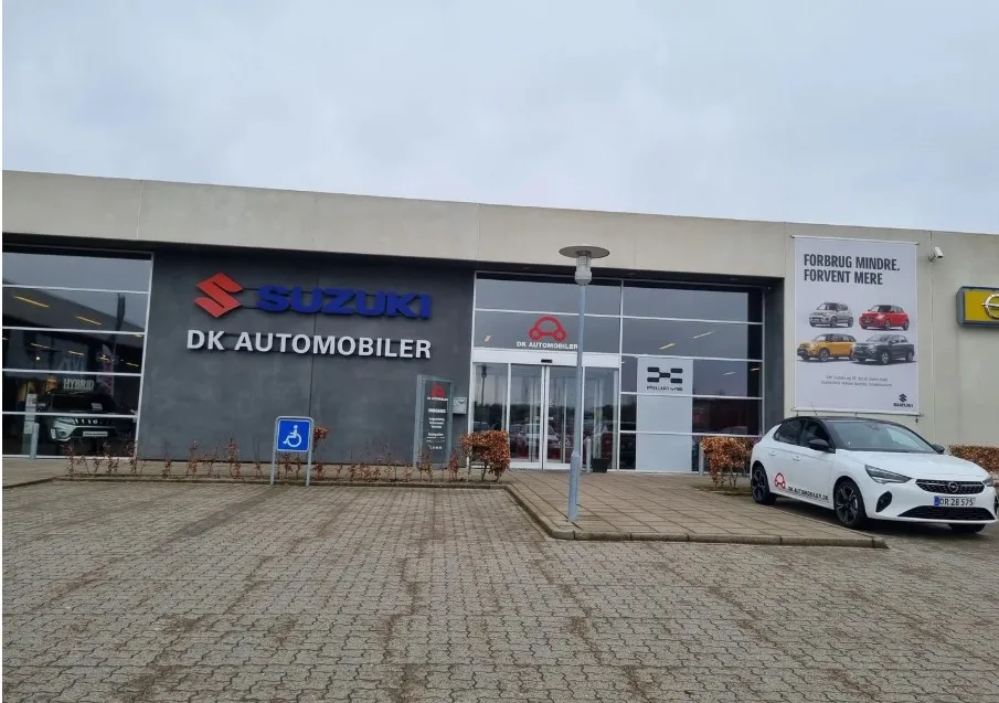 Suzuki DK Automobiler Vejle, med moderne showroom, en hvid bil parkeret og en banner med teksten Forbrug mindre, forvent mere