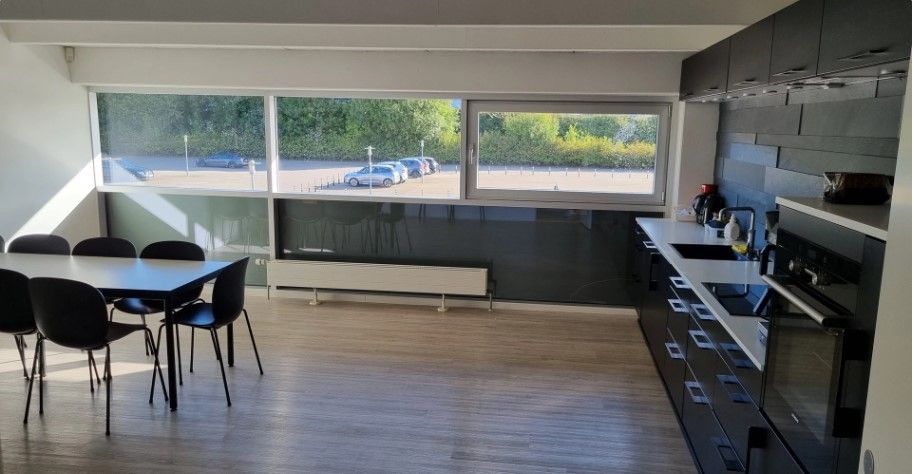 Lejlighed med moderne køkken, gult bord, sorte stole, radiator, vindue og udsigt til parkeringsplads med biler