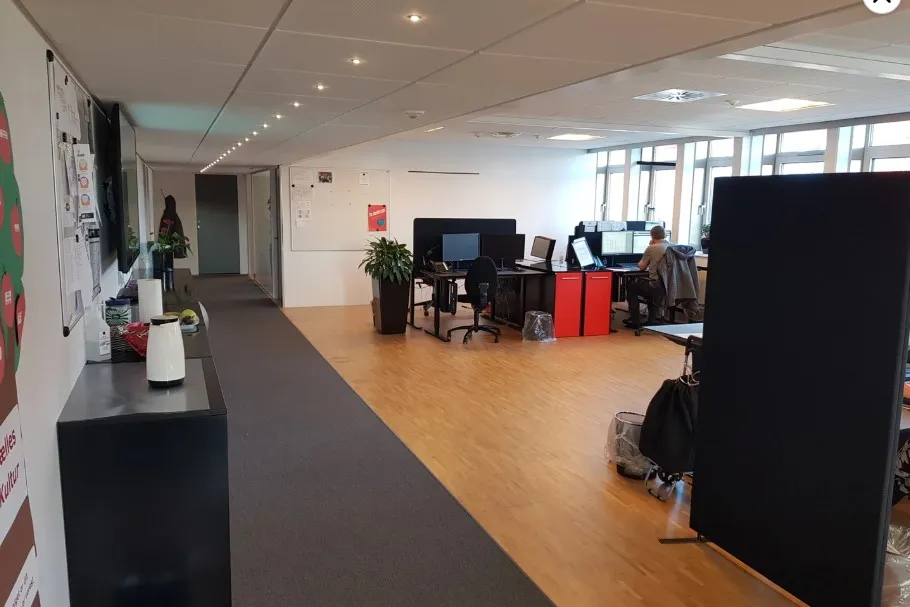 Kontor med moderne møbler og computere, hvor en person sidder og arbejder. Væggene er dekoreret med plakater og papirark.