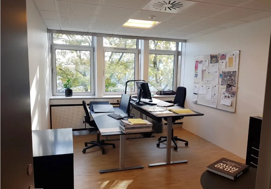 Kontor med skriveborde, computere, stol, plante og papirer samt en stor skiltstavle med noter og billeder