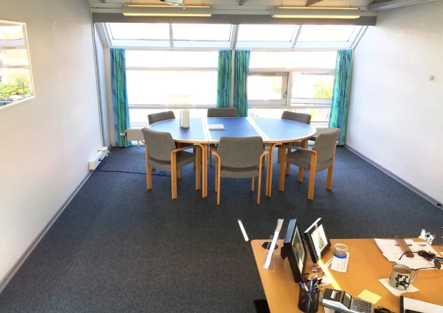 En stor mødesal med runde borde og stole, vinduer med gardiner og hvide vægge. Et skrivebord med computer, briller og papir