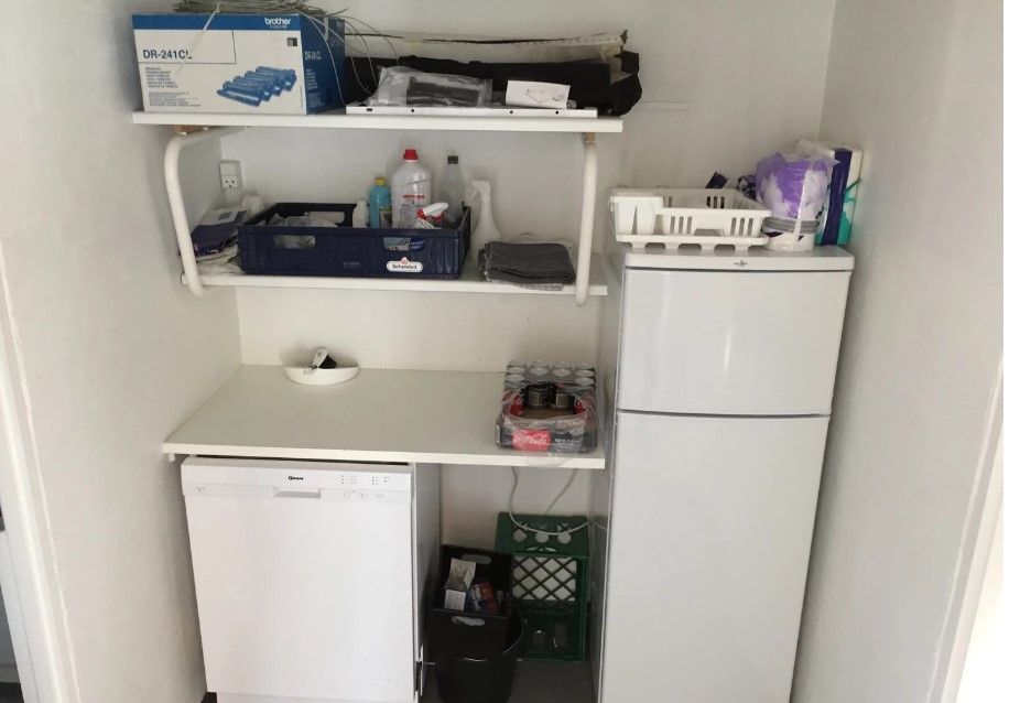 En lille lagerrum med to hvide hylder, en køleskab og en opvaskemaskine og en række kasser og flasker