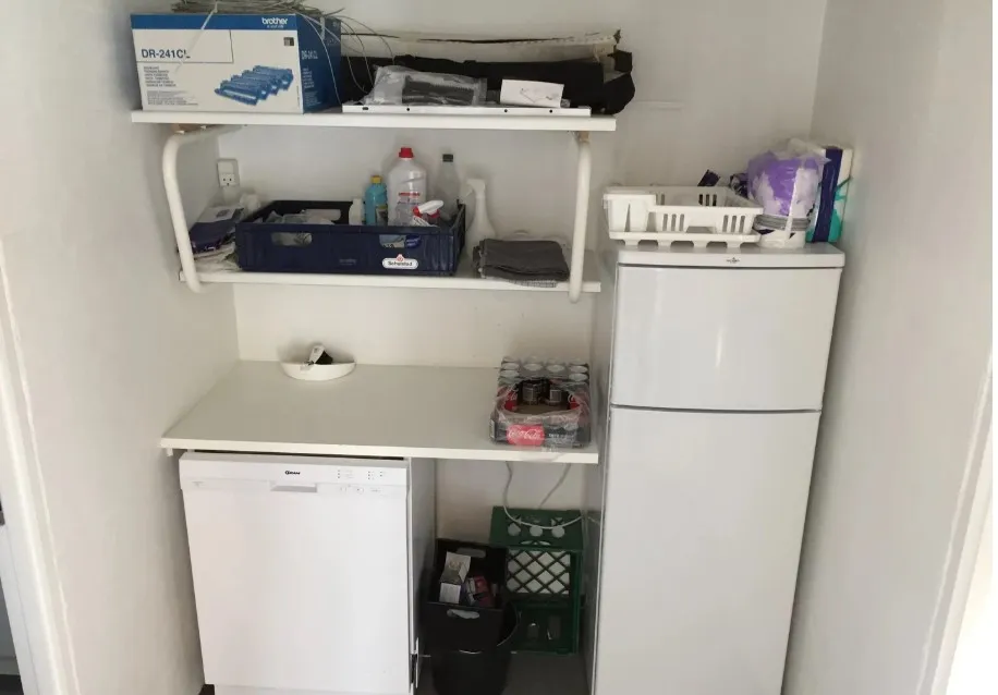 En lille lagerrum med to hvide hylder, en køleskab og en opvaskemaskine og en række kasser og flasker