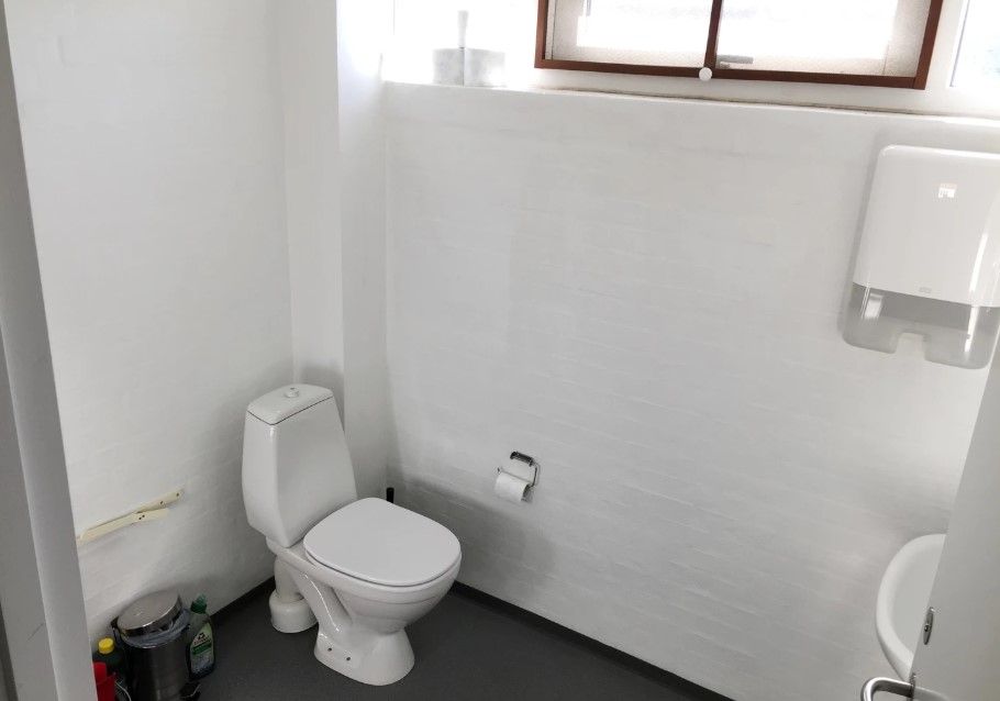 En toiletrum med en hvid toilet, håndsæbe dispenser, en toiletpapirholder og en hvid vandhaner