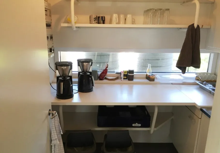 Et lille køkken med to kaffemaskiner og et skab med opbevaringsbokse og to papirkurve under bordet