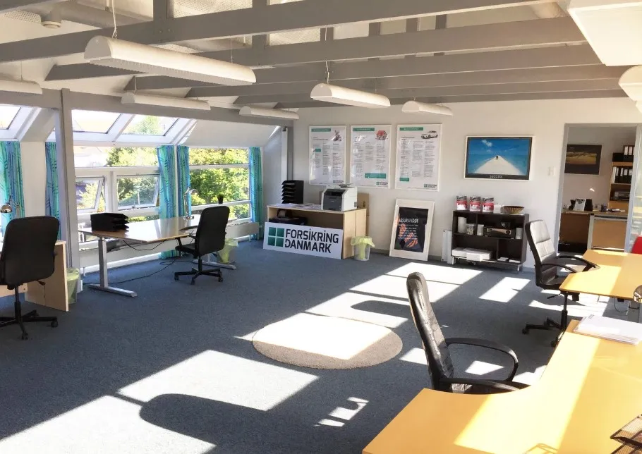 Et lyse kontor med bord, stol og papirkurv. På væggen hænger et billede og nogle plakater. Loftet er udstyret med hvide