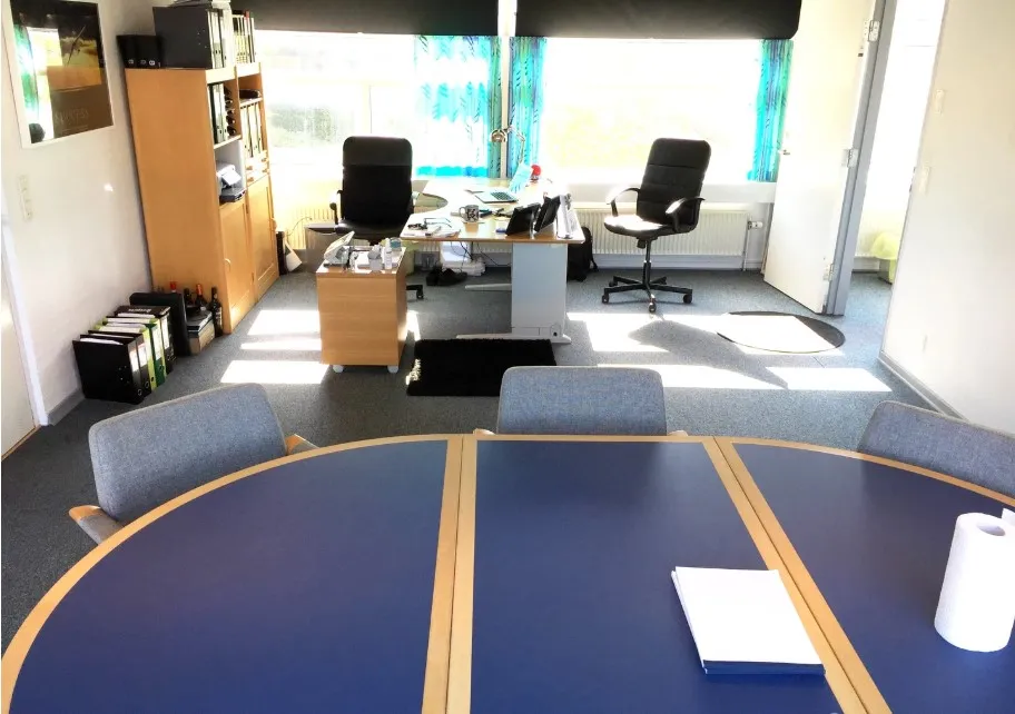 Et stort kontor med lyse gulve og vinduer. Bord og stole er spredt rundt. Der er et skrivebord med papir og en rulle