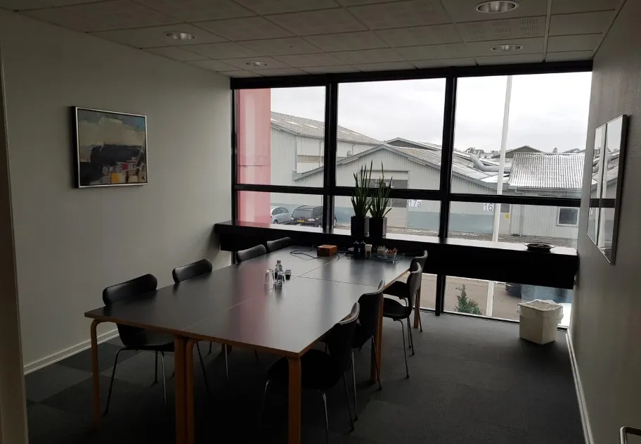 Kontor med bord og stole, gulvbelægning og vinduer med udsigt til bygninger og biler parkeret udenfor