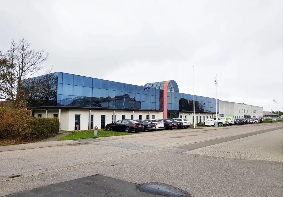 Store glasruder og høje vægge på bygningen, flere parkerede biler foran og et flag på en flagpæl