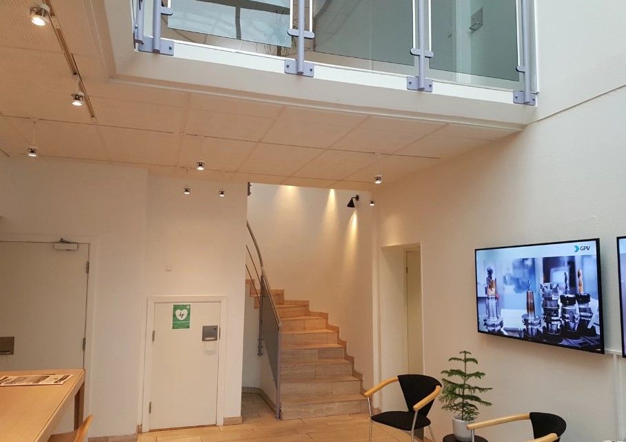 Inde i lokalet er der en hvid væg og en stor tv-skærm med teksten GPV. To stole med en plante og en bord er placeret på