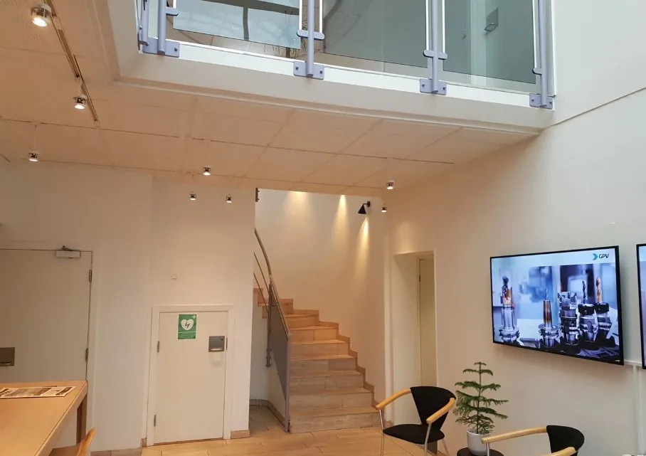 Inde i lokalet er der en hvid væg og en stor tv-skærm med teksten GPV. To stole med en plante og en bord er placeret på