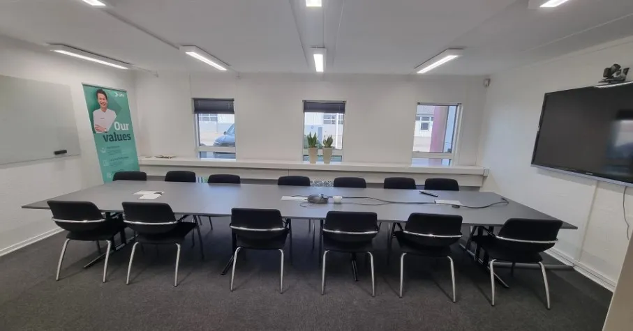 Store mødelokale med skrivebord, stole, whiteboard, projektor og plantepoter på vindueskarmen.