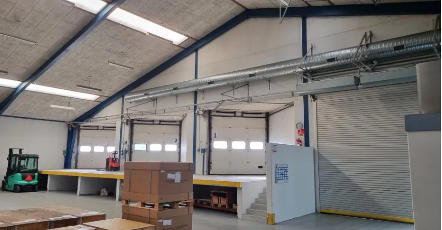 En stor lagerhal med flere åbne garageporte, flere lagerkartoner og en grøn gaffeltruck.