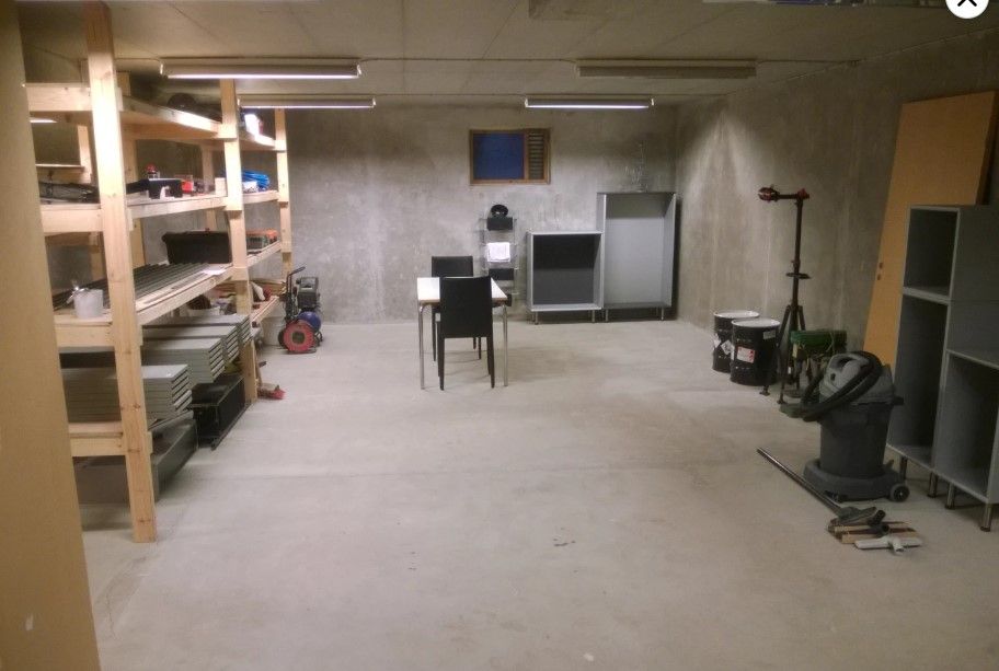 Et stort, tomt kælderrum med flere hylder, træstolper, en lille bordet med stole og en støvsuger.