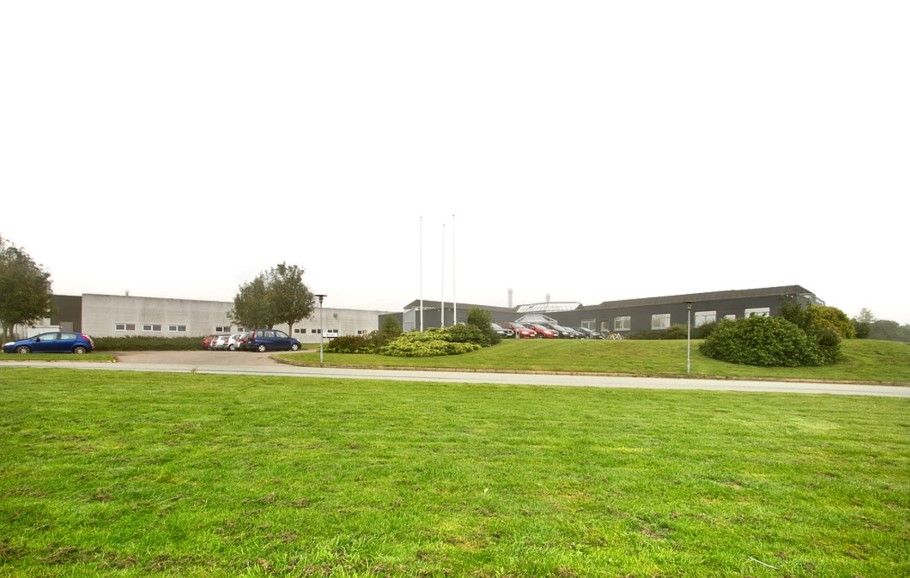 To biler parkeret på et grønt parkeringsområde med skrånende bakke og buske foran en bygning med mange vinduer