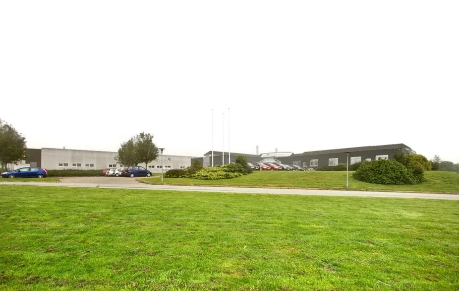 To biler parkeret på et grønt parkeringsområde med skrånende bakke og buske foran en bygning med mange vinduer