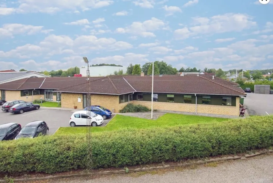 Kontorbygning med parkeringsplads og busstop foran. Flere biler parkeret. Busstop med bord og stol. Grønne planter og buske.