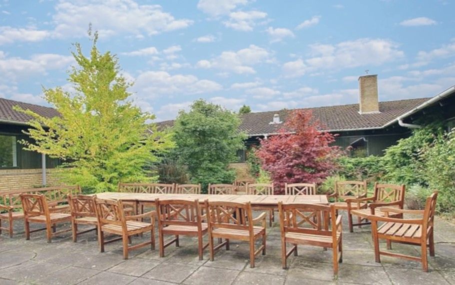Terrasse med træbord og stole til møder og sammenkomster med baggrund af kontorbygning og park.