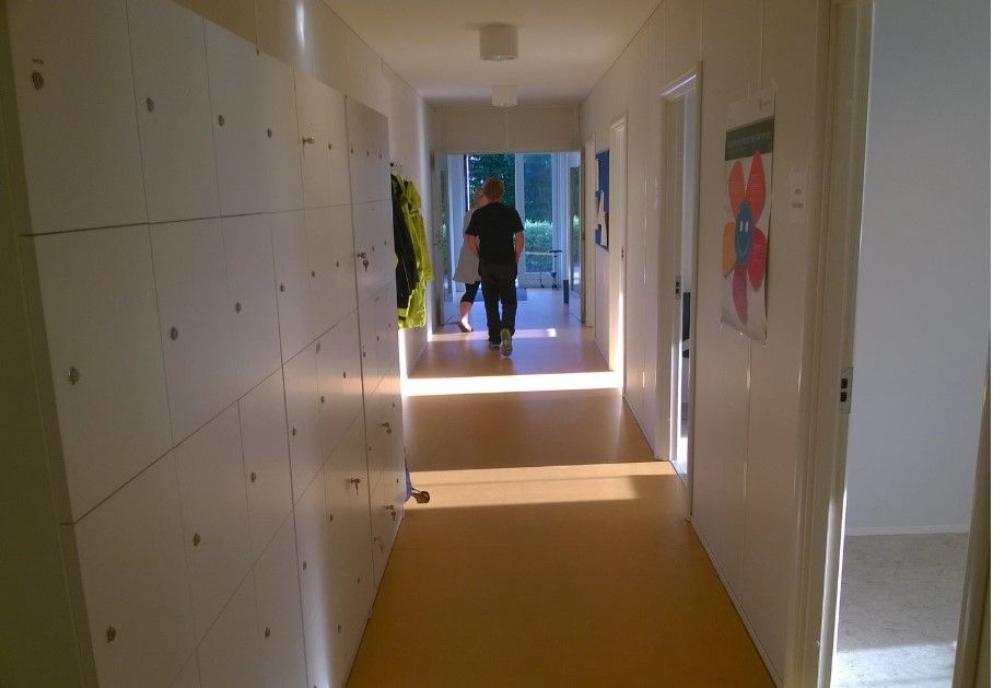 Hallen med hvide vægge og gulv, hvor en mand og en kvinde går. Der er hængeskabe og lysekroner i loftet og en væg med