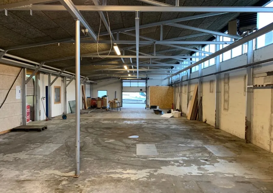 En tom industrihal med metalbærere, vinduer og træmateriale til loftet. Et hjul er parkeret ved indgangen.