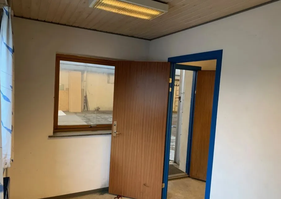 En tom industrilokale med lysegul væg og to døre i højre side. Der er et vindue med samme lysegul væg og en åben dør, der