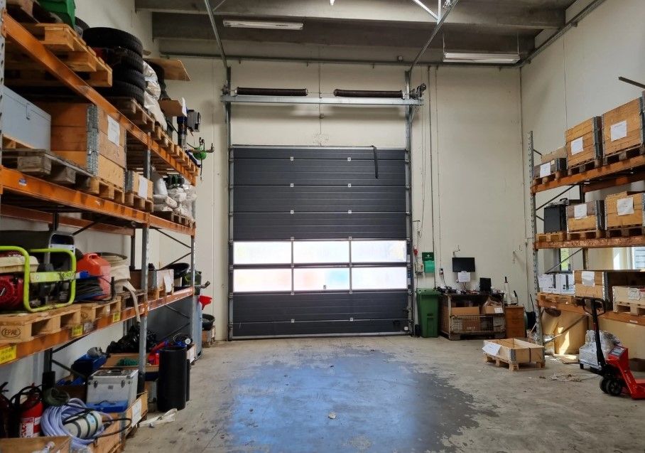 Lagerhal med store garagedøre, højloft, to rækker hylder, opbevaringsbokse, røde brandslukkere og træpaletter.