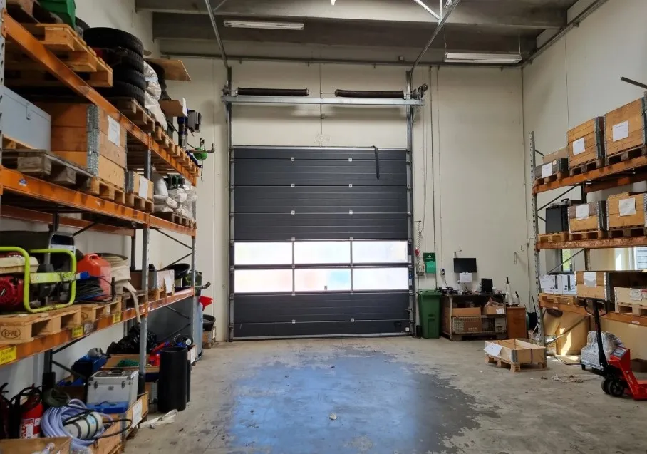 Lagerhal med store garagedøre, højloft, to rækker hylder, opbevaringsbokse, røde brandslukkere og træpaletter.