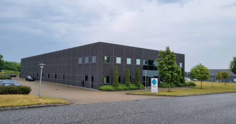 Lagerhal med søjler og store vinduer, parkeringsplads og biler, og grønne planter foran bygningen.