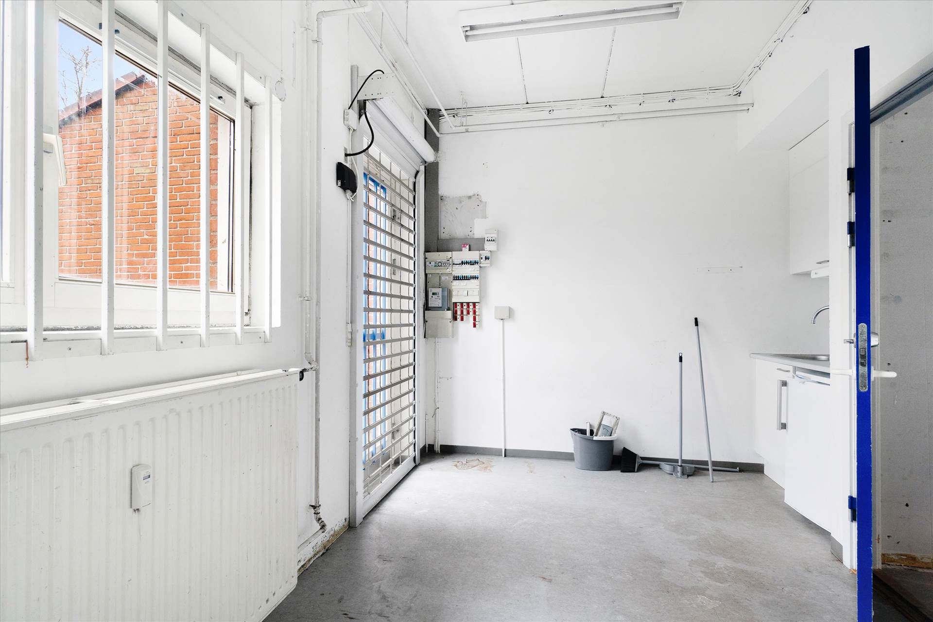 Værelse med vinduer og radiatorer samt dør med børster og skraldespand og apparater ved væggen