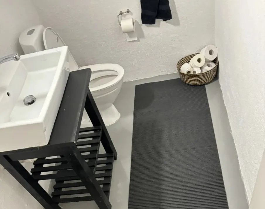Et lille badeværelse med hvid toilet, hvidt vasketøj, sort gulvmåtte og lysebrun kurv med toiletpapir