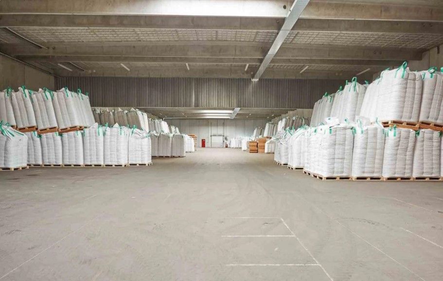 Lager med flere søjler med hvide beholdere og sække med grønt og hvidt bånd. Betongulv og lyse lofter.