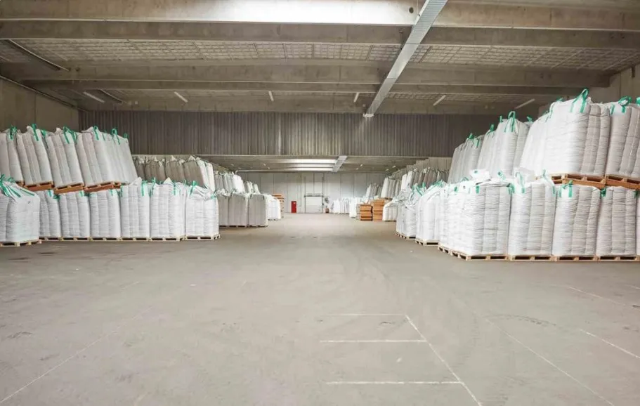 Lager med flere søjler med hvide beholdere og sække med grønt og hvidt bånd. Betongulv og lyse lofter.