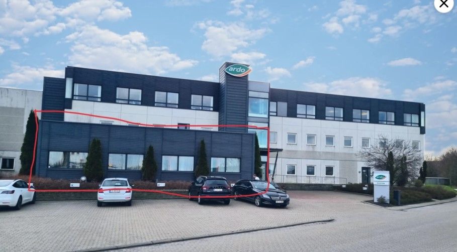 Kontorbyggeri med mange vinduer og parkeringsplads med fire parkerede biler foran og skov i baggrunden