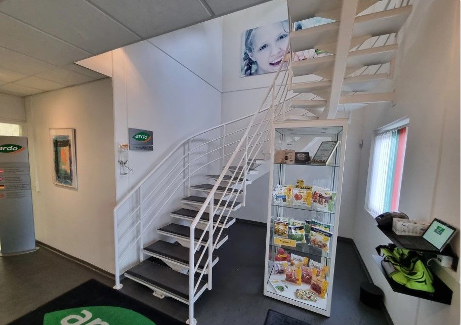 Kontorlokale med trapper, hvidt gulv, og vinduesvisning. På bordet står en laptop, en pose, og et display med snacks. Væggen