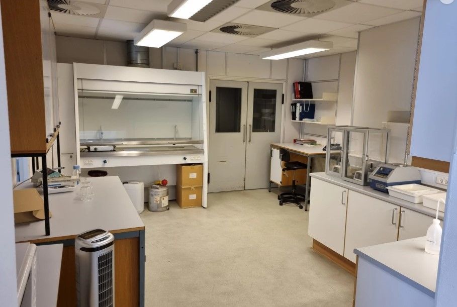 Lyst kontorlokale med hvide vægge og gul gulv. Dobbelt døre i midten og et apparat på arbejdsbordet. To stol og et bord med