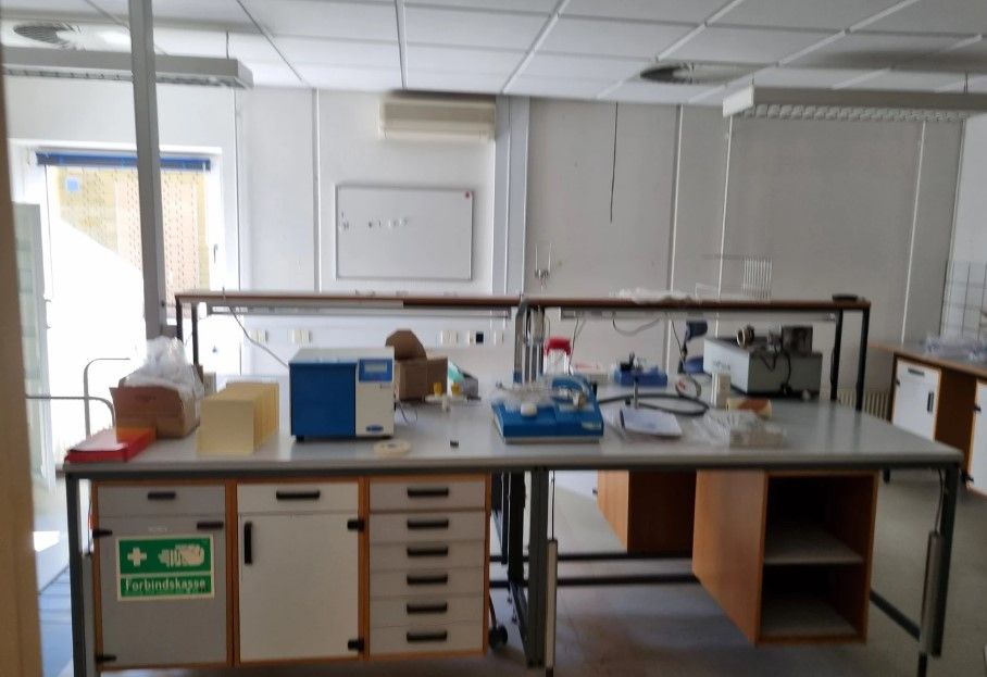 Laboratoriebord med diverse apparater og instrumenter, inklusiv stålborde, kasserne og hvidt pap, som er nyttige til