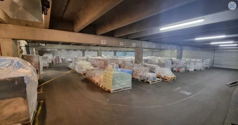 Mange lagerpæle med forskellige produkter pakket i plast på hver side. Pladsen er bred og er dækket af asfalt.