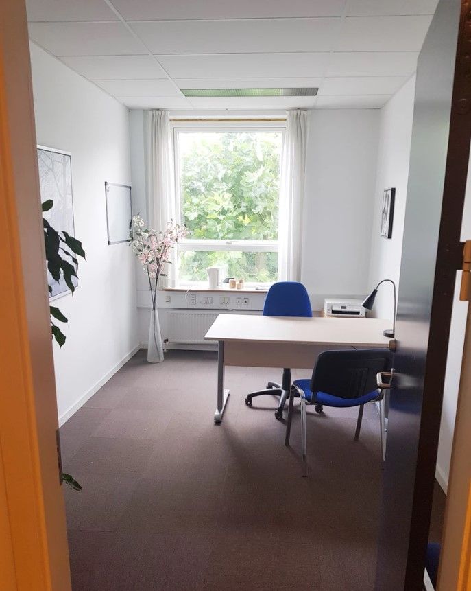 Et rent og lyse kontor med en hvid skrivebord og to blå stole med hjul. I hjørnet står en vase med lyserøde blomster. Ved