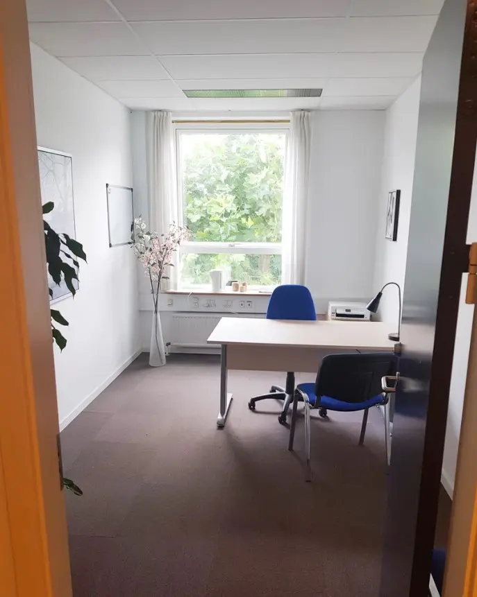 Et rent og lyse kontor med en hvid skrivebord og to blå stole med hjul. I hjørnet står en vase med lyserøde blomster. Ved