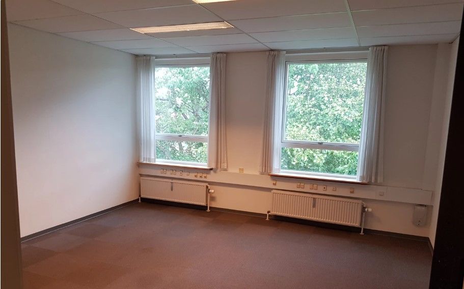 Et tomt kontor med hvide vægge, højt til loftet og trævinduer med hvide gardiner og et radiatorpar.