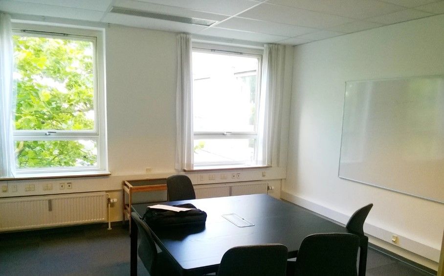 Et tomt kontor med tre stole, et bord med en taske og en tavle på væggen. Der er to vinduer med hvide gardiner, der stiger