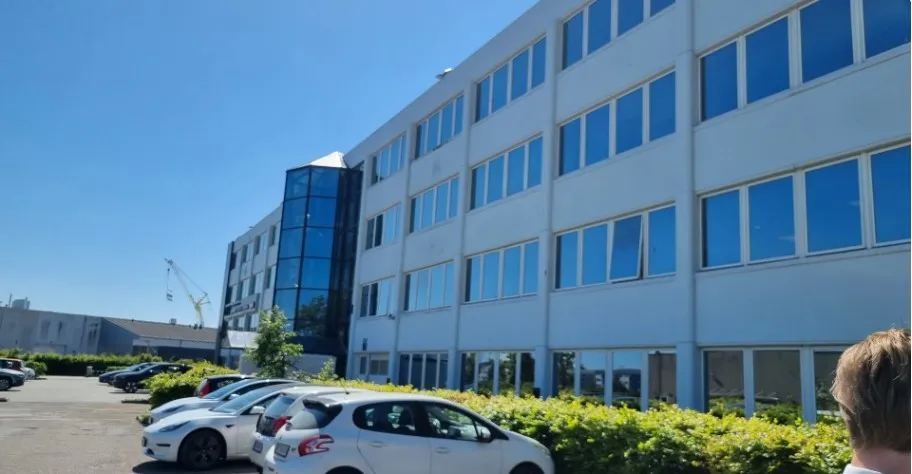 En stor, hvid bygning med glasruder og kontorer i flere etager med parkeringsplads og biler parkeret foran