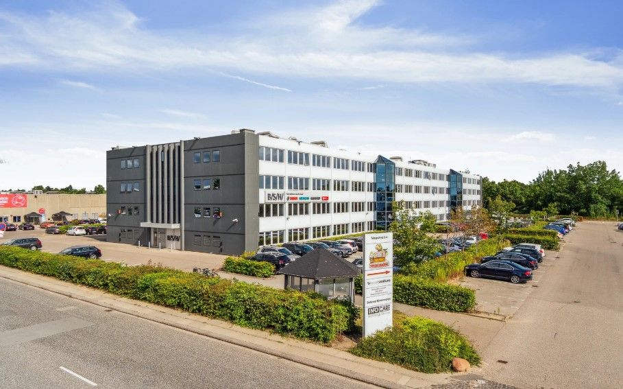 Kontorbyggeri med glasvinduer og busseltræer. Parkerede biler og grønne buske i forgrunden. Infoskilt med navn og logo