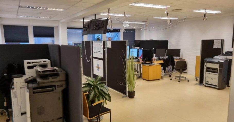 Kontorlokale med moderne møbler, printere, og plante. Lyse loftslampe og monitorer i kontoret.