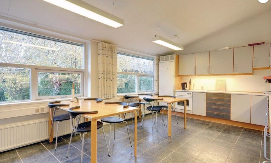 En tom møde- og kontorværelse med lyse gulvfliser, lyse loftslampe, to lange træborde med høje stole samt en stor køleskab