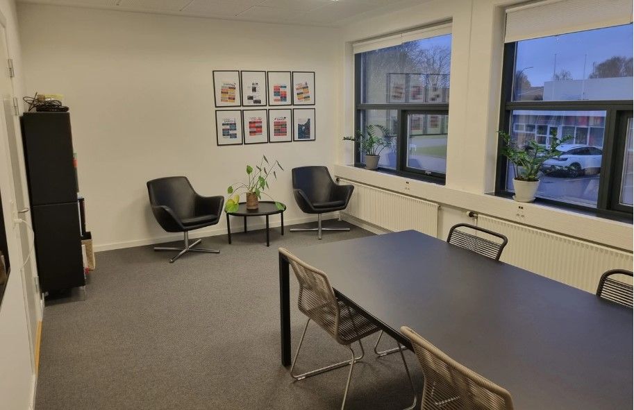 Kontorlokale med lyse, tavle og plante. Fire stole og et bord er placeret i rummet.