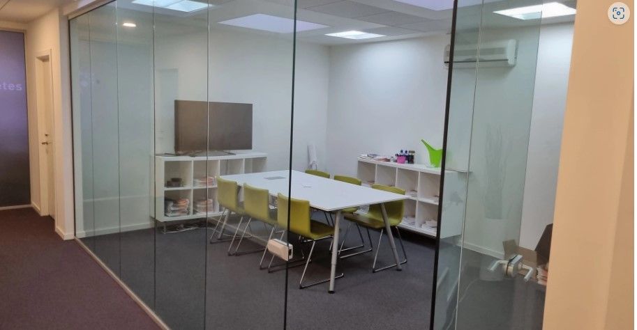 Kontorlokale med glasvægge, hvidt bord og stole, tv og højtalere, samt to hvide hylder med diverse genstande