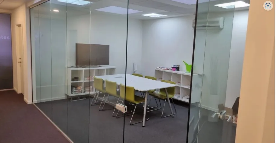 Kontorlokale med glasvægge, hvidt bord og stole, tv og højtalere, samt to hvide hylder med diverse genstande
