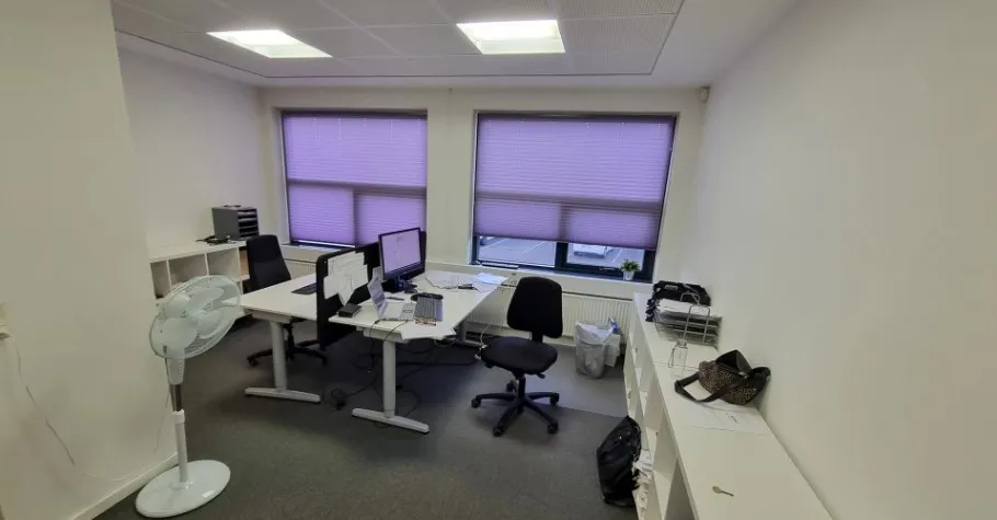 Kontor med højloft, vinduer med jalousier, to skriveborde, stol, fan, skraldespand og plante i vindueskarmen